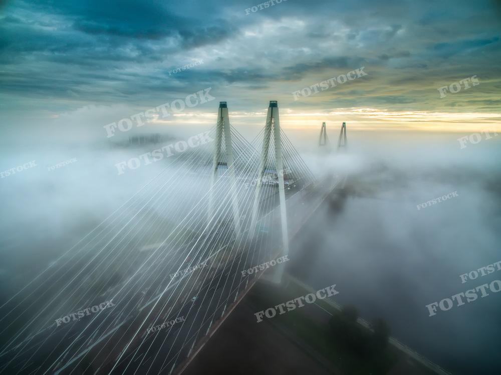 Эта загадочная фотография демонстрирует мост, частично скрытый в облаках тумана. Игра света и тени, а также мягкий свет, пробивающийся сквозь облака, создают мистическую и впечатляющую атмосферу. Такой снимок может быть использован для иллюстрации статей или блогов о необычных погодных условиях, в качестве фона для вдохновляющих цитат или мотивационных плакатов, а также для добавления уникальной атмосферы в книги или фильмы, где требуется создать ощущение таинственности или изоляции.